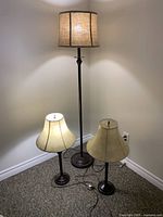 Three lamps shown cornered against walls, two table lamps with cream fabric shades, and one floor lamp with linen-look drum shade, all with metal bases.