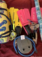 Two pairs of flippers (yellow and pink), two snorkels with masks, gray foot pump, and rolled beach mat with flip-flop prints. All laid out on a red surface.