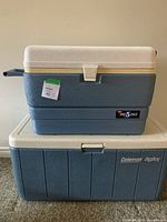 Two hard-sided coolers stacked: Coleman Big Boy on bottom and Igloo Max 5 Cold on top. Both have blue bodies and white lids with latch locks and handles.