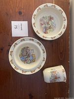 Three ceramic Bunnykins dishware pieces on wooden tabletop: two shallow bowls and one cup, all featuring Bunnykins bunny illustrations and rabbit border.