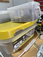Photo showing plastic containers stacked, filled with various screws and bolts, including a yellow-lidded container.