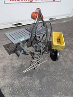 Overview of lot showing 2 grey foldable plastic tables, metal wheel-barrel planter with yellow container, metal hanging plant stands, horseshoes, and stakes on pavement.