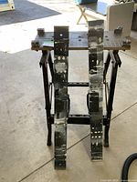 Foldable work table with clamping surface and two black metal collapsible saw horses in front, showing paint residue and used condition.