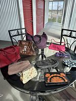 All 10 assorted handbags and purses arranged on a black glass table with black chairs around, showing variety in color and style.
