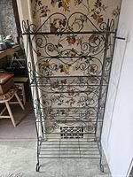 Front view of the wire metal bakers rack showing decorative scrollwork, four shelves, and black metal wire construction against floral wallpaper background.
