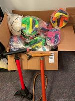 Box filled with 30 new beach volleyballs in colorful designs, each wrapped in plastic, alongside two hand air pumps.