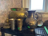 Showing the entire lot including the hammered brass planter, miniature log holder, two Indian brass cups, Italian brass vase, and metal candleholder on a black rack shelf near a window.