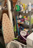 Wide view of basement area showing ironing board, shelving unit with cleaning supplies, mop, vacuum, laundry basket and hangers.