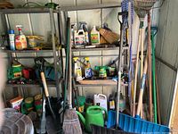 Full view of metal shelves with garden chemicals, fertilizer bottles, brooms, and various garden tools leaning against the back wall of shed
