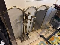 Fireplace tools set displaying four tools topped with duck-head designs on a stand with matching gold finish, placed in front of fireplace screen.