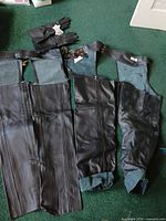 Photo of two pairs of black genuine leather chaps – one new with tags size small and one worn size XL – alongside a pair of new with tags black leather gloves, placed on green carpet.