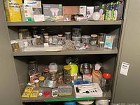 Full shelf view with rows of jars containing assorted fasteners and packages of light bulbs on the top shelf. Miscellaneous hardware and tools on lower shelf.
