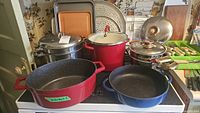 Multiple vintage pots include a red cast iron casserole pot, blue cast iron pot, stainless steel pots and lids, placed on a stovetop.