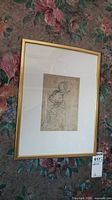 Full view of gold framed print with sketch of kneeling woman, lying on floral carpet.