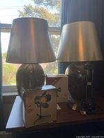 Pair of matching large table lamps with dark elliptical bases and metallic silver conical shades on a wooden table in front of a window. Two boxed studio and clamp lamps are visible in front.