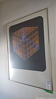 Angled photo of framed geometric cube cluster poster showing 3D cube blocks in orange and purple on black background, with white mat and simple frame.