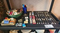 Full assortment on shelf showing bowl and boxes of vintage matchbooks, glass with plastic stir sticks, and collector spoons laid out. Includes lot tag.