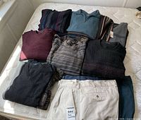 Lot containing seven men's sweaters in various colors and styles along with three pairs of denim pants and one striped scarf, shown on a mattress.