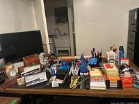Wide view of the desk showing many office supplies including pens, sticky notes, letter openers, keyboards, lamps, and a monitor.
