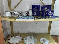 Top shelf showing various china teacups with floral designs, Selangor pewter boxed items behind, Wedgwood plate, glass pickle tray, and porcelain plates on lower shelf