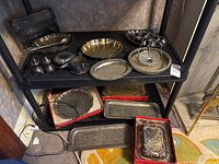 Two black shelves holding an assortment of silver plated serving pieces including trays, covered dish, salt and pepper shakers, salad servers, and napkin rings.