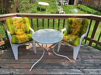 Two white plastic armchairs with thick floral patterned seat cushions in yellow and green, placed next to a round white plastic outdoor table with metal legs.