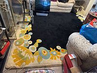 Area rug shown in living room setting with furniture around it, featuring thick black pile and floral border detail.