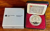 Coin displayed in open black clamshell box with outer silver Royal Canadian Mint box on wood surface.