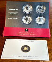 Four sterling silver 50 cent coins in maroon plastic display case with black insert. Coins are encapsulated and arranged in two rows with certificate booklet below.