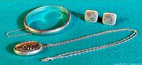 Overview image showing the sterling silver locket necklace, the English silver clasp bangle with engraved pattern and safety chain, and the pair of square silver screw back earrings.