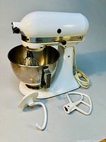 Full side view of KitchenAid mixer with stainless steel bowl and three attachments next to it: dough hook, flat beater, wire whisk.