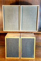 Front view of two pairs of bookshelf speakers: top pair has beige cloth covers with teak wood frames; bottom pair has black cloth covers with lighter wood frames.