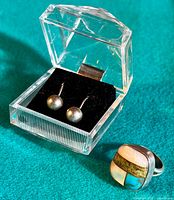 Earrings displayed in a transparent box with black insert and ring shown next to it on green fabric background.
