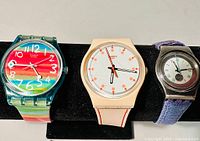 Three vintage Swatch watches displayed on a black stand showing the distinct colors and designs of each watch.