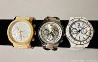Three watches in a row on black display bar: gold-tone, silver-tone, white resin.