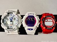 Full view of the three watches side by side, showing the white G-Shock, white Baby-G with purple face, and red translucent Baby-G watch.