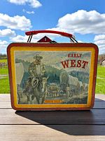 Front view of 1980s Early West metal lunchbox. Shows lithographed illustration of cowboy on horseback and covered wagons with forest and mountain background. Red plastic handle attached on top. Good lighting and outdoor background.