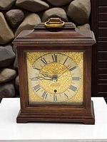 Front view of the Seth Thomas mantel clock showing the brass-tone ornate clock face with Roman numerals, black hands including second hand, and wood case with brass handle on top. Clock face has intricate floral embossing.