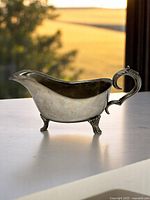 Side view of silver-plated vintage gravy boat showing curved spout, scrolled handle and floral cast feet, placed on a table with a natural outdoor background.