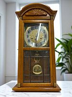 Front view of the ERGO mechanical pendulum clock showing wood case, glass panels with grid detail, ornate clock face with black numerals, and brass pendulum behind lower glass door.