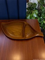 Front angled view showing the leaf-shaped teak tray placed on a wooden surface with visible natural grain and smooth finish, highlighting the organic shape and division into two sections.