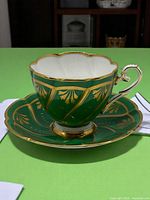 Teacup and saucer set displayed on green cloth background, showing gold leaf design and gilded handle.