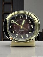 Front view of Westclox Baby Ben alarm clock showing black sunburst dial with gold numerals, luminous hands, and gold-tone bezel mounted on cream plastic casing with base.