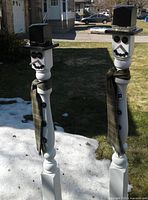Pair of tall painted cedar post snowmen with black hats and scarves standing outdoors on patchy snow and grass, rebar bases visible at ground level.
