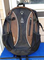 Front view of medium size black and brown Rambler backpack by Woods brand showing logo, bungee cord, and front zipper pockets.