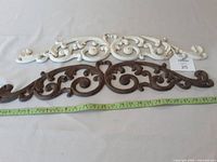 Two cast iron architectural design features, one white and one dark brown, both ornamental with scroll designs, laid flat with a measuring tape showing length approximately 24 inches.