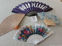 Four handheld folding fans laid flat: purple with 'QUEEN PLEASE!', beige with cut-outs, blue floral, lace floral fan