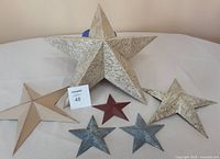 Photo of six decorative metal stars in various colours and sizes arranged on a white surface against a white wall. Includes 1 large beige star (14"), 2 medium stars (11") in beige and red, and 3 small stars (5") in metallic grey and red.
