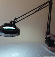 Side view of the black magnifying lamp attached to an adjustable arm held by a person, showing the circular magnifier with light inside.