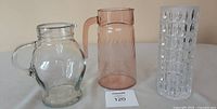 Three glass items shown together on surface against plain background: clear glass pitcher, pink Depression glass pitcher, cut crystal vase.